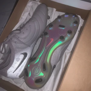 NIKE TIEMPO LEGEND X ELITE HELT NYA OANVÄNDA - Snygga grå Nike fotbollsskor i syntetmaterial med en cool, skimrande sula. De har snörning och en svart Nike-logga på sidan. Perfekta för fotbollsspelare som vill ha både stil och funktion på planen.