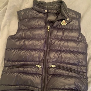 Mörkblå gui väst från Moncler - Säljer en snygg mörkblå dunväst från Moncler i mycket bra skick. Västen har en dragkedja framtill och två praktiska fickor med dragkedjor. Perfekt för höst och vår när du vill hålla dig varm men ändå se stilren ut. Passar både till vardags och fest! Men den har en liten defekt på ryggen men som knappt syns vilket jag har förseglat med tyg tejp i liknande färg (bild 4) OBS skriv till mig innan ni köper via plick!