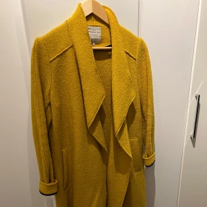 Gul kappa från Urban Code - Säljer en superfin gul kappa från Urban Code. Den är lång och har en snygg, draperad krage. Perfekt för höst och vår! Materialet känns mjukt och lite texturerat, vilket ger en cool look. Ärmarna är långa och kan vikas upp för en mer avslappnad stil. Passar perfekt till både vardag och fest!