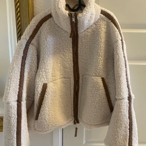 Ny cremevit teddyjacka storlek S från H&M - Ny med tags. Supermysig vit pilejacka med bruna detaljer och dragkedja framtill. Perfekt för kyliga dagar med sin fluffiga och varma design. Jackan har en hög krage och två fickor för extra komfort. En stilren och trendig jacka som passar perfekt till höst och vinter. Den är lätt vadderad  storlek S (oversize)