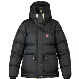 Svart dunjacka från Fjällräven - Säljer en svart dunjacka från Fjällräven i ganska bra skick. Jackan har långa ärmar och en praktisk huva. Den är perfekt för kalla vinterdagar och har både dragkedja och knappar för extra skydd mot kylan. Fjällrävens logga syns på bröstet. Passar perfekt för vintern! Tygbiten som dragkedjan sitter på sitter lite löst längst ner men inget som stör funktion eller syns! (Se sista bilden)