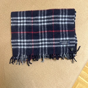 Rutig halsduk från Burberry - Säljer en klassisk rutig halsduk från Burberry i mörkblått, grått och rött. Den är gjord av 100% lammull och har fransar i ändarna. Perfekt för att hålla sig varm och stilren under kyliga dagar. Tillverkad i Skottland och i mycket bra skick!