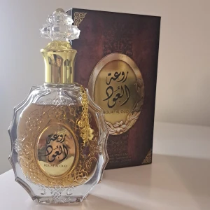Rouat Al Oud, EDP, från Lattafa - Säljer en lyxig parfym från Lattafa, Rouat Al Oud. Flaskan är elegant med en gyllene kork och detaljer, och har en vacker, genomskinlig design med ett guldfärgat emblem. Doften är förmodligen rik och orientalisk, perfekt för speciella tillfällen. Kommer i en snygg förpackning som matchar flaskans stil. Perfekt för dig som älskar exklusiva dofter!