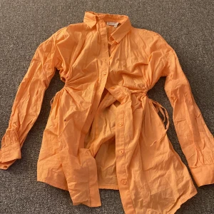 Orange skjortklänning med knytband - Säljer en snygg orange skjortklänning med knytband i midjan. Den har långa ärmar och knappar framtill. Perfekt för vår och sommar! 🧡