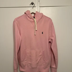 Rosa hoodie från Ralph Lauren - Säljer en rosa hoodie från Ralph Lauren i bra skick. Den har en liten logga på bröstet och en mysig känguruficka. Det finns ett litet hål på ärmen, men annars är den i fint skick. Perfekt för en avslappnad stil!