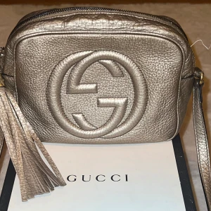 Guldig axelväska från Gucci - Säljer en snygg guldig axelväska från Gucci i mycket bra skick. Väskan har en elegant fransdetalj och ett stort GG-emblem på framsidan. Perfekt för både vardag och fest! ✨
