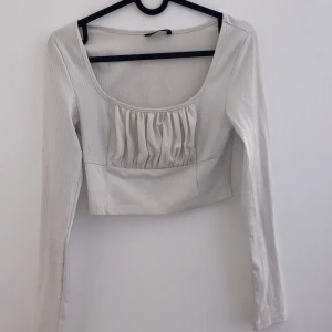 Vit långärmad topp - Säljer en stilren vit topp med långa ärmar och en fin rynkad detalj framtill. Perfekt för både vardag och festliga tillfällen. Toppen har en tight passform och är gjord i ett mjukt material som känns skönt mot huden.