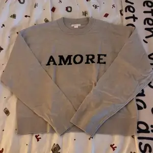 Säljer en snygg beige stickad tröja från Gina Tricot med texten 'AMORE' i svart på framsidan. Tröjan har långa ärmar och en bekväm passform. Perfekt för en avslappnad stil under hösten eller våren. 🧡