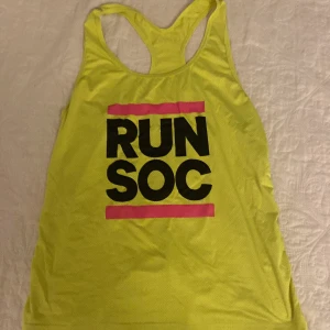 Gul linnetopp från Stadium - Säljer en sportig gul linnetopp från Stadium med texten 'RUN SOC' i svart och rosa. Perfekt för träning! Tillverkad i polyester och elastan för en bekväm passform. Toppen är ärmlös och har en racerback-design.