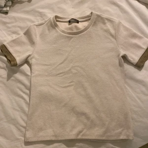 Vit t-shirt från Zara - Säljer en stilren vit t-shirt från Zara i storlek XS. Den har korta ärmar med snygga guldfärgade detaljer vid ärmsluten. Perfekt för både vardag och festliga tillfällen. Materialet känns mjukt och bekvämt mot huden. Passar perfekt till jeans eller kjol!