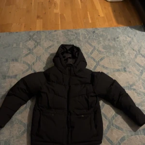 Svart pufferjacka med huva - Säljer en svart pufferjacka i mycket bra skick. Den har en skön huva och praktiska fickor med knappar. Perfekt för kalla vinterdagar och vinterpromenader. Jackan är långärmad och har en dragkedja framtill. Passar både till vardags och lite mer uppklädda tillfällen. Har använt den två gånger 