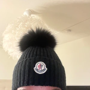 Svart ribbad mössa från Moncler - Säljer en snygg svart ribbad mössa från Moncler med en stor fluffig boll på toppen. Den är perfekt för kalla dagar och har en broderad Moncler-logga framtill. Mössan är i mycket bra skick och passar perfekt till vinterns alla outfits.