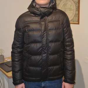 Svart pufferjacka från Calvin Klein - Säljer en snygg svart pufferjacka från Calvin Klein. Jackan är perfekt för vintern! Den har en dragkedja framtill och en mysig huva. Perfekt för att hålla sig varm och stilren under kalla dagar. 