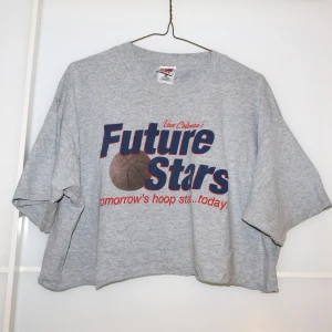 Grå croppad t-shirt från Fruit of the Loom - Säljer en grå croppad t-shirt från Fruit of the Loom i storlek XL. Den har ett tryck med texten 'Future Stars' och en basketboll. Perfekt för en avslappnad stil eller sportig look. T-shirten är i bomull och har korta ärmar.