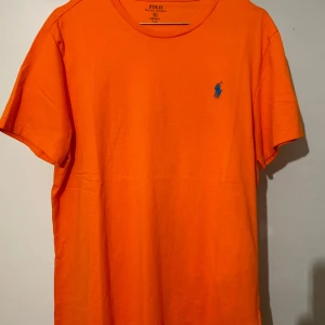 Orange t-shirt från Ralph Lauren - Säljer en snygg orange t-shirt från Ralph Lauren i bomull. Den har en klassisk passform med korta ärmar och en blå logga på bröstet. Perfekt för en avslappnad stil eller sommarens äventyr! 🧡