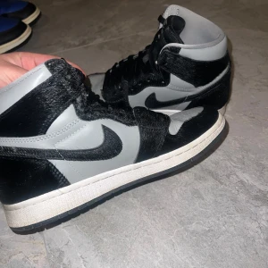 Air Jordans 1 Mid i svart och grått - Säljer ett par Nike Air Jordans 1 Mid i svart och grått i storlek 39. Skorna har en unik design med svart pälsdetalj. 