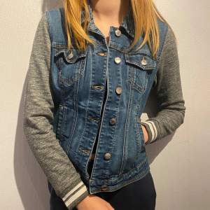 Snygg jeansjacka från Guess med grå ärmar i sweatshirt-material. Jackan har en klassisk jeansblå färg med knappar framtill och två bröstfickor och två framfickor med knappar. Perfekt för en casual look under vår och höst!