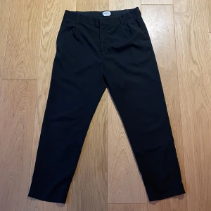 Svarta kostymbyxor från Weekday - Säljer ett par stilrena svarta kostymbyxor från Weekday. De är en aning vidare i passformen ner till. Perfekta för både formella tillfällen och vardagsbruk. Byxorna är i bra skick och har en enkel stängning med dragkedja och knapp.   Skick 9/10.