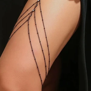 Svart benkedja med strassdetaljer - Snygg och unik svart benkedja som verkligen sticker ut! Den har flera tunna kedjor som hänger ner och är dekorerad med glittrande strassdetaljer. Perfekt för att ge din outfit en extra edge, kanske till en fest eller en speciell kväll.