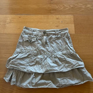 Beige kjol från Gallop - Säljer en superfin beige kjol från Gallop! Den har en cool volangdetalj och broderade mönster som ger den en unik look. Kjolen har också praktiska fickor med knappar.