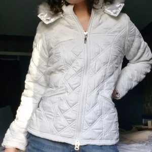 Vit quiltad jacka med fuskpäls från Ann Christine - Säljer en superfin vit jacka från Ann Christine. Den har en mysig huva med grå fuskpäls - perfekt för en vintrig y2k-stil! Jackan är i bra skick och redo för nya äventyr! Accepterar rimliga erbjudanden :)