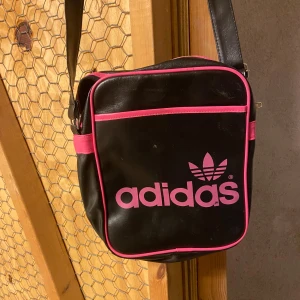 Svart och rosa axelväska från Adidas - Säljer en cool svart axelväska från Adidas med rosa detaljer och logga. Väskan har en justerbar axelrem och en praktisk dragkedja upptill. Perfekt för att bära med sig det viktigaste när du är på språng. Lite slitage syns på ytan, men den är fortfarande i bra skick.