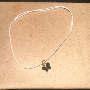 Armband med fjärilsberlock - Säljer ett enkelt och stilrent armband med en söt fjärilsberlock. Armbandet har en tunn vit tråd och berlocken är i svart och guld. Perfekt för att lägga till en subtil touch av elegans till din outfit!