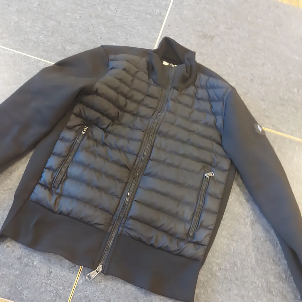 Säljer en snygg svart jacka från Moncler. Den har en quiltad design på framsidan och är utrustad med dragkedjor på fickorna. Jackan har långa ärmar och en cool logga på ärmen. Perfekt för höst och vår!. Jackor.