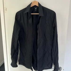 Svart skjorta från H&M - Säljer en stilren svart skjorta från H&M. Den är långärmad och perfekt för både vardag och fest. Skjortan har en klassisk krage och knappar hela vägen ner. Materialet känns som bomull, vilket gör den bekväm att bära. 