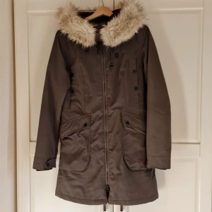 Parkas - Jacka i parkasmodell med avtagbar pälskrage. Två bröstfickor och stora fickor fram. Färgen är grågrön. Knappt använd.