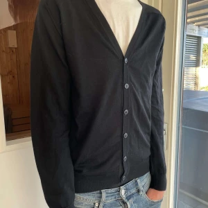 Svart kofta från Oscar Jacobson - Säljer en stilren svart kofta från Oscar Jacobson i storlek M. Den har en klassisk v-ringning och knappar framtill. Perfekt för både vardag och festliga tillfällen. Passar bra till jeans eller chinos för en avslappnad look.