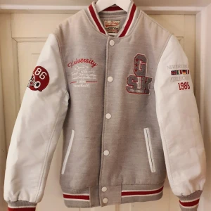 Baseballjacka - Baseballjacka/ collegejacka i grått (50% ull) med vita ärmar i syntetskinn. Snygga applikationer och broderier i retro/vintagestil, se bilder! Jackan är perfekt för höst och vår då den har ett mjukt och värmande foder. Mycket bra skick, dock har den blivit lite lätt blekt på axlarna. Storlek 14 år, ca 158 cm.