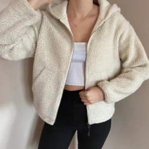 Beige teddyjacka med dragkedja - Mysig beige teddyjacka med dragkedja framtill. Perfekt för kyliga dagar med sin fluffiga och varma design. Jackan har en avslappnad passform och är långärmad, vilket gör den idealisk för höst och vinter.