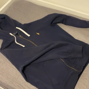 Ralph lauren  - Säljer en snygg mörkblå hoodie från Ralph Lauren med dragkedja och vit snörning i huvan. Den har en liten gul logga på bröstet och är perfekt för en avslappnad stil. Passar bra till både jeans och joggers.