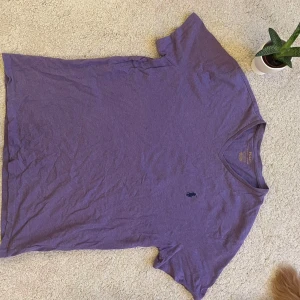 Lila t-shirt från Ralph Lauren - Säljer en snygg lila t-shirt från Ralph Lauren med v-ringning och broderad logga på bröstet. Perfekt för en avslappnad stil. Tillverkad i mjukt material för extra komfort.