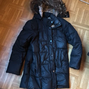 Svart tjock dunjacka burberry - Säljer en stilren svart dunjacka från Burberry med klassiskt rutigt foder. Jackan har en avtagbar huva med mjuk bra fuskpäls och stängs med både dragkedja och knappar. Perfekt för kalla vinterdagar. Vet inte im den är äkta lr inte. Då jag själv i second hand. Kom inte till användning här då jag har en liknande. Därav låga priset.