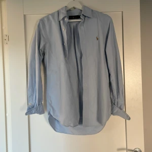 Blå skjorta från Ralph Lauren - Snygg ljusblå skjorta från Ralph Lauren med klassisk krage och broderad logga på bröstet. Skjortan har långa ärmar och knappar framtill. Perfekt för en stilren look. Finns tyvärr lite missfärgning på skjortan, kolla bild fyra o fem. Bild 4 är från insidan o bild fem från utsidan! Priset kan diskuteras! Mvh 