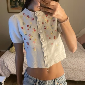 Vit kortärmad kofta med blommor från & other storys - Söt vit kofta med korta ärmar och färgglada blommor broderade på framsidan. Den har knappar hela vägen och en croppad passform. Perfekt för en somrig look!