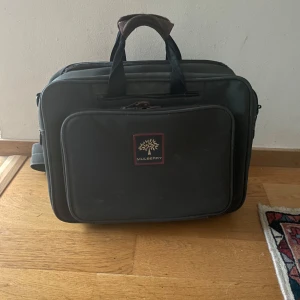 Datorväska från Mulberry - Stilren svart/grå datorväska från Mulberry. Väskan har en praktisk axelrem och flera fack med dragkedja. Klassisk design. Har i det andra facket 1 av 2 trasig dragkedja, samt att kedjan är sliten. Går fortfarande att stänga och öppna