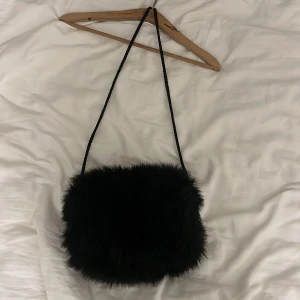 Svart pälsväska med axelrem - Snygg svart axelväska i pälsmaterial. Perfekt för att ge din outfit en trendig touch. Väskan har en enkel design med en lång axelrem för bekväm användning.