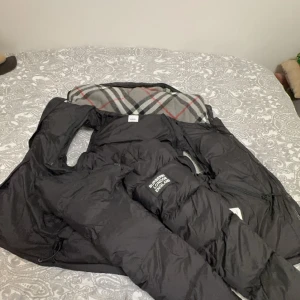 Burberry jacka/väst - Säljer en snygg svart dunjacka från Burberry med ett klassiskt rutigt foder. Jackan har en dragkedja och långa ärmar, perfekt för kyliga dagar. Den är både stilren och funktionell. Kan göras till väst också. Finns i Malmö