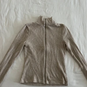 Beige ribbad tröja med dragkedja - Säljer en stilren beige ribbad tröja med dragkedja och hög krage. Tröjan har långa ärmar och är perfekt för kyligare dagar. Passar bra till både jeans och kjol.