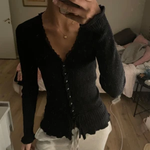 Svart ribbad kofta med knappar - Snygg svart ribbad kofta med knappar framtill och knytband i midjan. Perfekt för kyligare dagar och kan enkelt matchas med olika outfits. Den har långa ärmar och en v-ringad design.