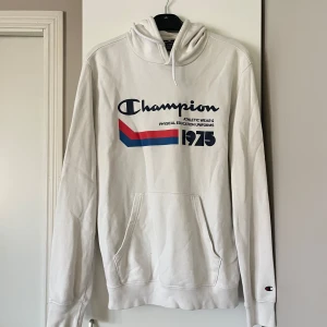 Vit hoodie från Champion - Vit hoodie från Champion med tryck i blått och rött. Hoodien har en känguruficka och justerbar huva med snören. 