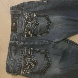 Y2k Miss Me Jeans Bootcut - Snygga blå jeans från Miss Me med dekorativa bakfickor och glittriga detaljer. Jeansen har en bootcut-stil och är perfekta för en trendig look. De har knappar, diamanter och dragkedja framtill.  Hade gärna velat behålla de här fina Jeansen som är i jättebra skick men går tyvärr inte eftersom att de är för stora för mig och är storleken 29.