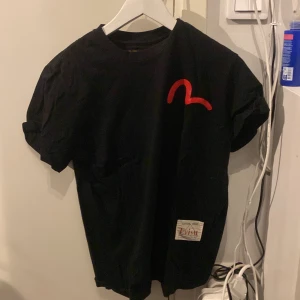 Svart t-shirt från Evisu med tryck - Snygg svart t-shirt från Evisu med ett stort färgglatt tryck på baksidan som föreställer en katt och texten 'Mighty Daruma'. Framsidan har en röd logotyp. Perfekt för dig som gillar unika och iögonfallande plagg!