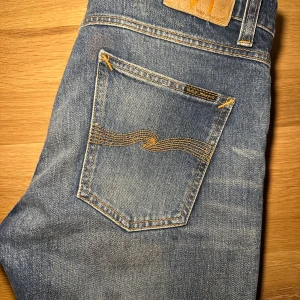  Nudie Jeans - Säljer ett par klassiska blå jeans från Nudie Jeans med snygga slitningar och kontrastsömmar. De har en normal passform och är perfekta för en avslappnad stil. Modellen är lean dean och sitter något större än vanliga jeans