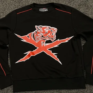 Svart tröja från Plein Sport - Snygg svart tröja från Plein Sport med ett coolt rött tigertryck på framsidan. Tröjan har långa ärmar och röda detaljer längs axlarna och ärmarna. Perfekt för en sportig look!