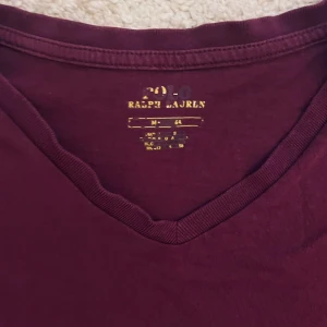 Vinröd t-shirt från Ralph Lauren - Säljer en vinröd t-shirt från Ralph Lauren med v-ringning och broderad logga på bröstet. Perfekt för en avslappnad stil. T-shirten är kortärmad och gjord i mjukt bomullsmaterial.