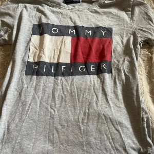 Grå t-shirt från Tommy Hilfiger - Säljer en grå t-shirt från Tommy Hilfiger med det ikoniska logotyptrycket i rött, vitt och svart på framsidan. Perfekt för en avslappnad stil.